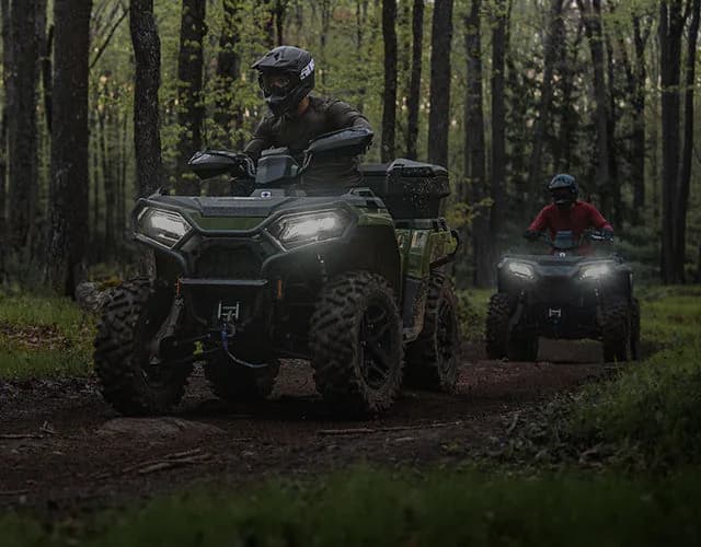 Two ATVs riding a wooded trail with headlights on